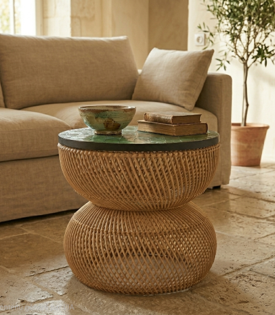 Woven rattan two-tier side table with a green top, a ceramic bowl, and two old books, placed beside a beige sofa in a sunlit living room.